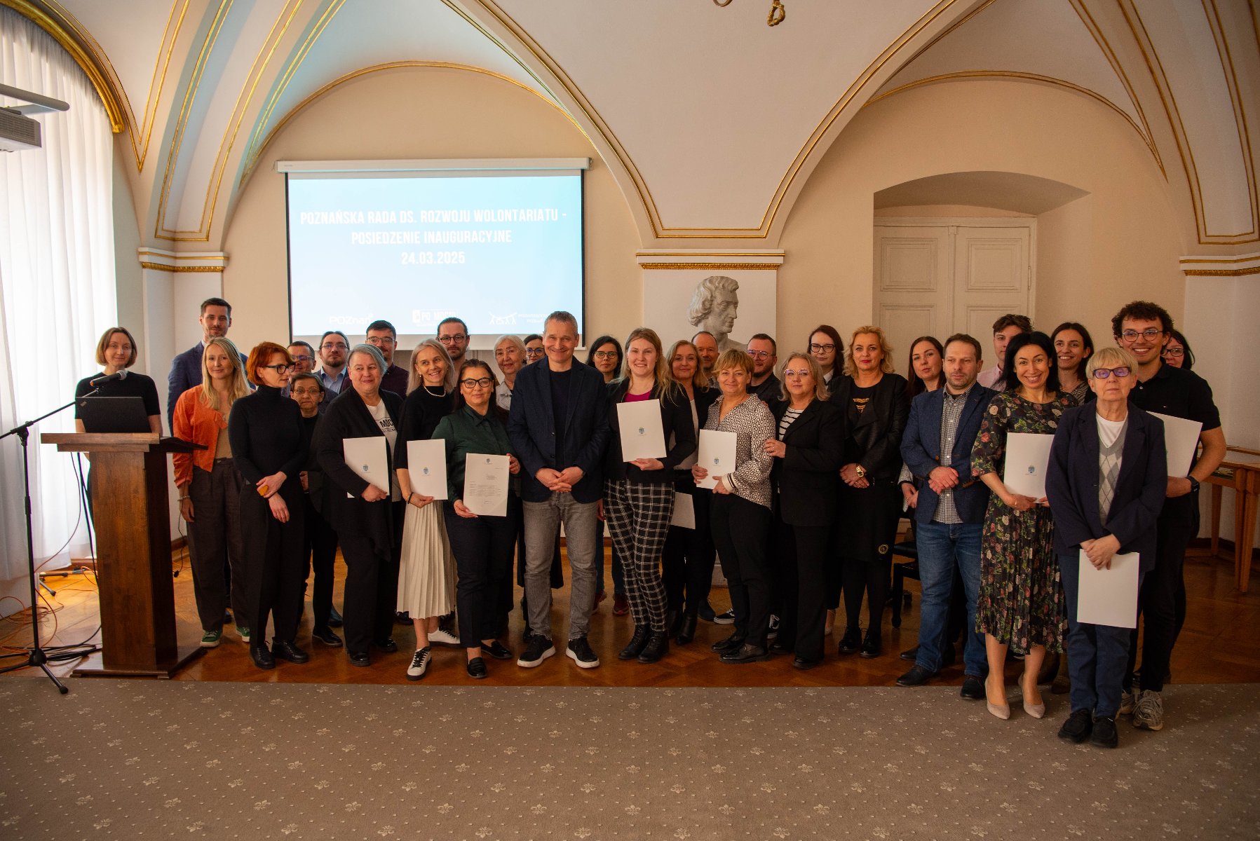 Grupa ludzi w eleganckim wnętrzu z łukowym sufitem i bogatymi dekoracjami. Osoby pozują do zdjęcia przed ekranem z napisem: ,POZNAŃSKA RADA DS. ROZWOJU WOLONTARIATU - POSIEDZENIE INAUGURACYJNE 24.03.2025'. Po lewej stronie podium z mikrofonem. Część osób trzyma certyfikaty lub dokumenty.