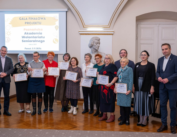 Grupa osób pozuje do zdjęcia w sali z drewnianą podłogą, podczas Gali Finałowej. W centrum ujęcia w rzędzie stoi dziesięć starszych kobiet, trzymających certyfikaty, z uśmiechem skierowanym w stronę aparatu.