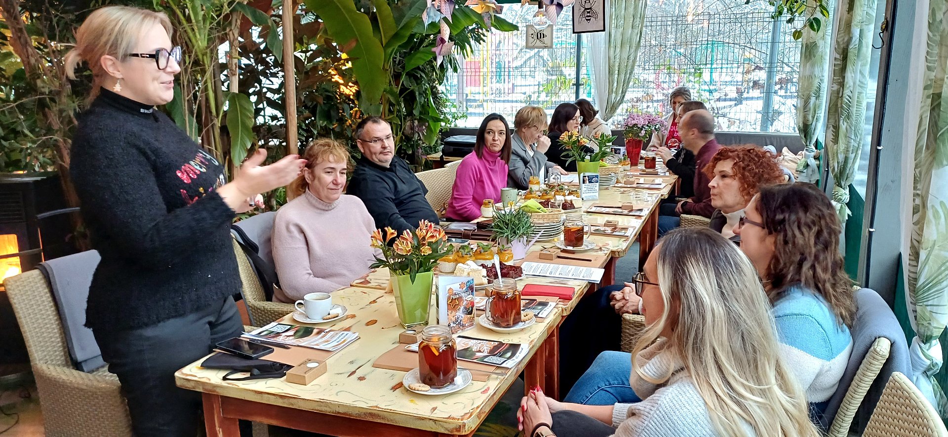 Grupa osób siedzi przy długim jasnym stole, na stole znajdują się filiżanki, talerzyki, ciasto i notatniki oraz ulotki, z lewej strony stoi blondynka w okularach i opowiada coś reszcie zgromadzonych przy stole osób