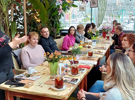 Grupa osób siedzi przy długim jasnym stole, na stole znajdują się filiżanki, talerzyki, ciasto i notatniki oraz ulotki, z lewej strony stoi blondynka w okularach i opowiada coś reszcie zgromadzonych przy stole osób