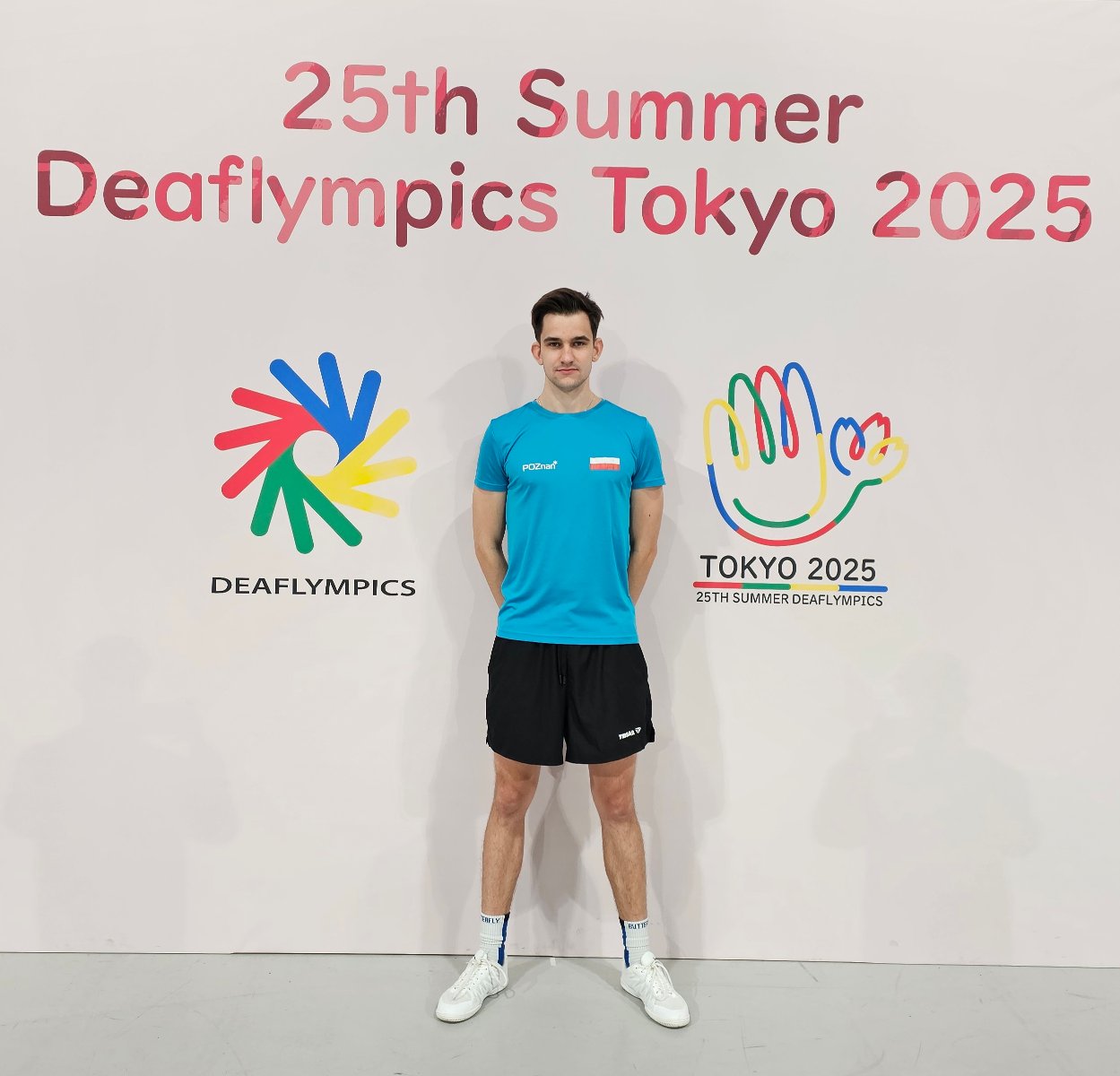 Grzegorz Nowak na XXV Letnich Igrzyskach Głuchych - zawodnik stoi w stroju sportowym na tle ścianki zawodów