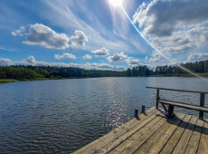 Jezioro z kawałkiem pomostu, w tle znajduje się brzeg z drzewami. Pogoda jest słoneczna, woda niebieska.