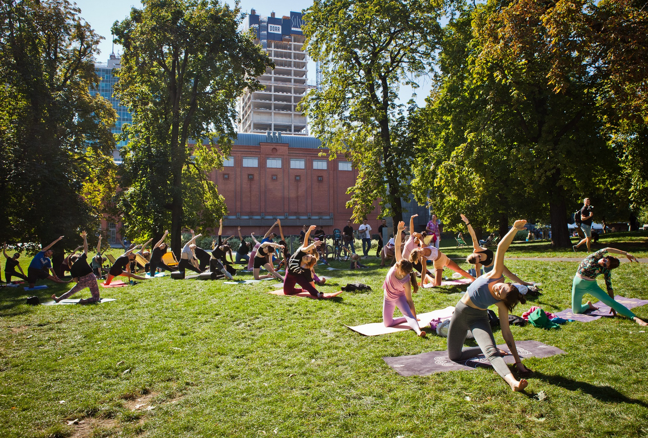 Zdjęcie parku przy Strarym Browarze. Na trawie, pod drzewami jogę uprawia grupa osób.