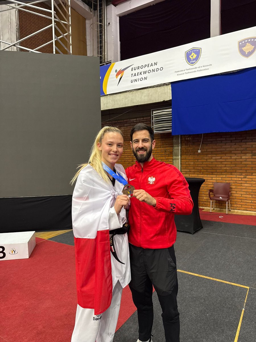 Julia Nowak z brązowym medalem ME U-21 w taekwondo, fot. Facebook Julia Nowak Taekwondo Olimpijskie - na zdjęciu uśmiechnięci kobieta i mężczyzna. Kobieta jet owinięta flagą i ma na szyi medal, a mężczyzna trzyma ten medal i ma strój sportowy w barwach narodowych