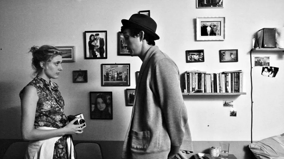 Czarno-białe zdjęcie mężczyzny i kobiety stojących przodem do siebie i rozmawijających w salonie. Za nimi ściana z fotografiami i kanapa.