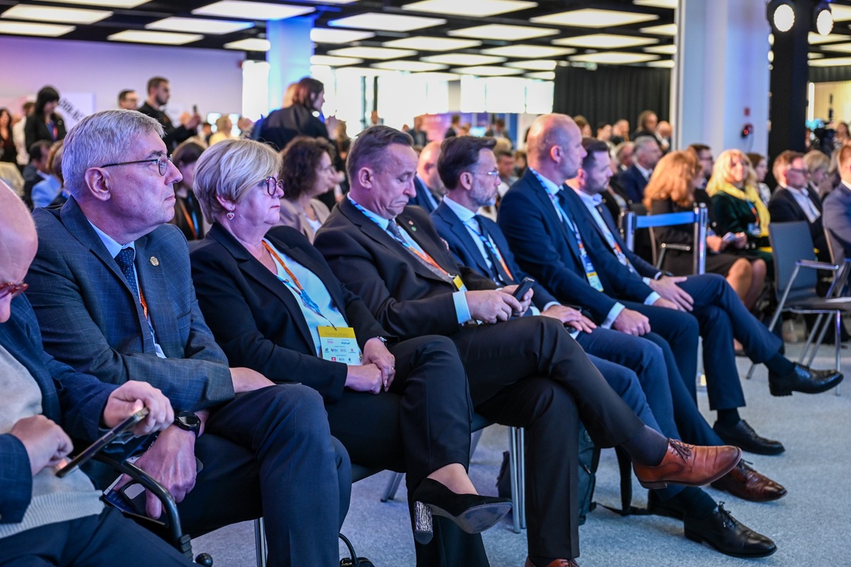 kilka osób siedzi pierwszym w rzędzie na krzesłach w sali konferencyjnej, za nimi kolejne rzędy krzeseł i osób