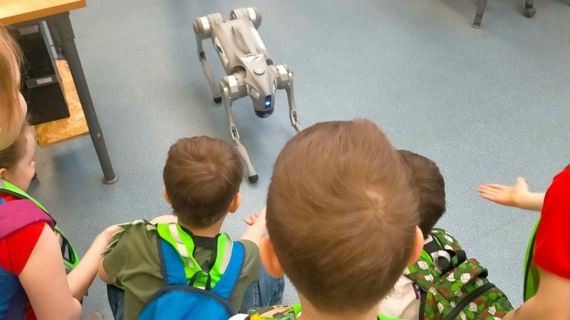 Kilkoro dzieci siedzi na podłodze i obserwuje czworonożnego robota przypominającego psa w szkolnej sali.