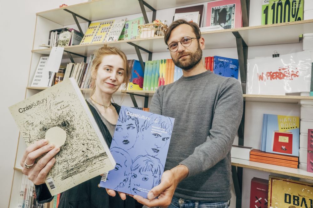 Kobieta i mężczyzna stoją na tle regału prezentując egzemplarze czasopisma.