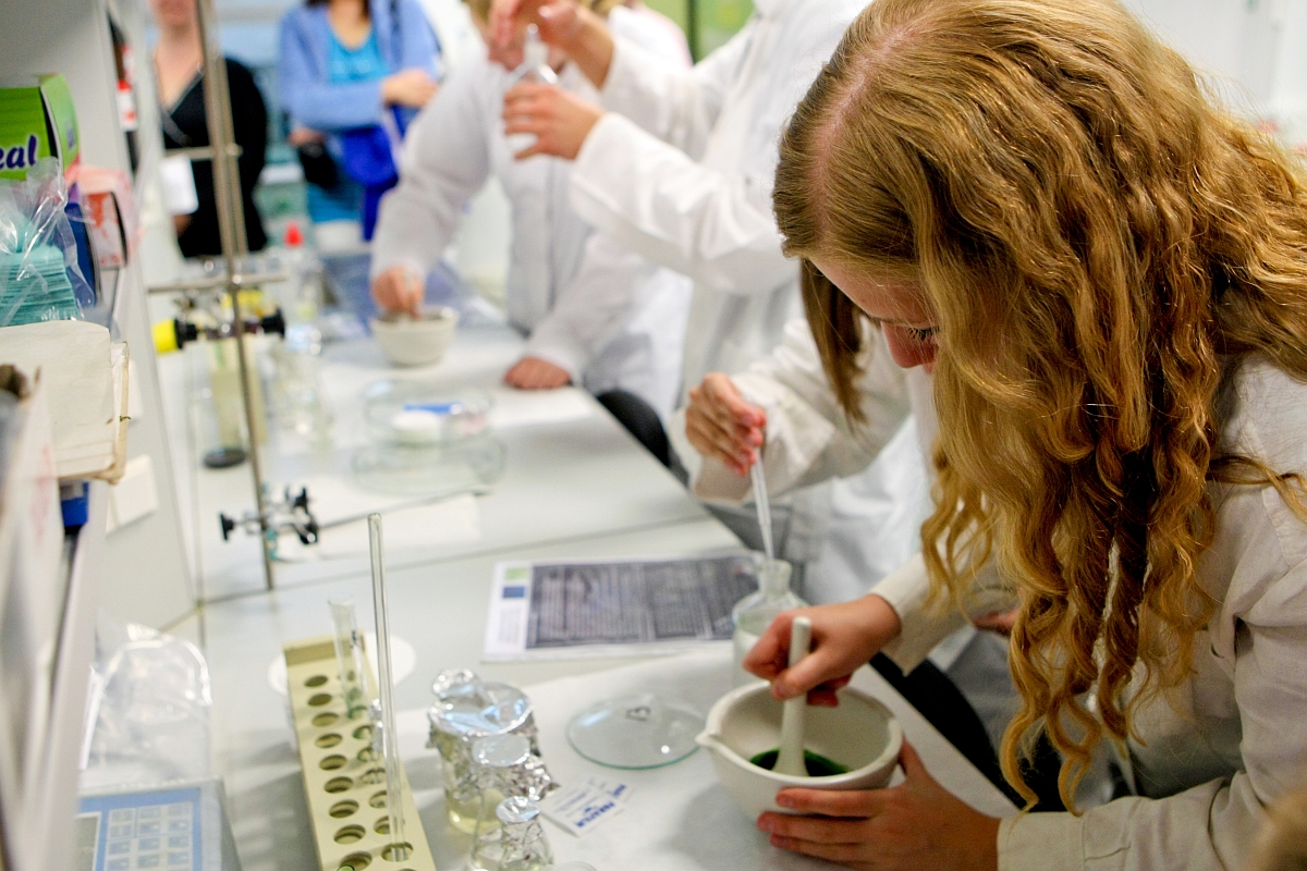 Kobieta w długach falowanych blond włosach w białym kitlu pochyla się nad stołem w laboratorium, miesza zielony płyn w małej białej misce. W tle niewyraźnie widać inne osoby