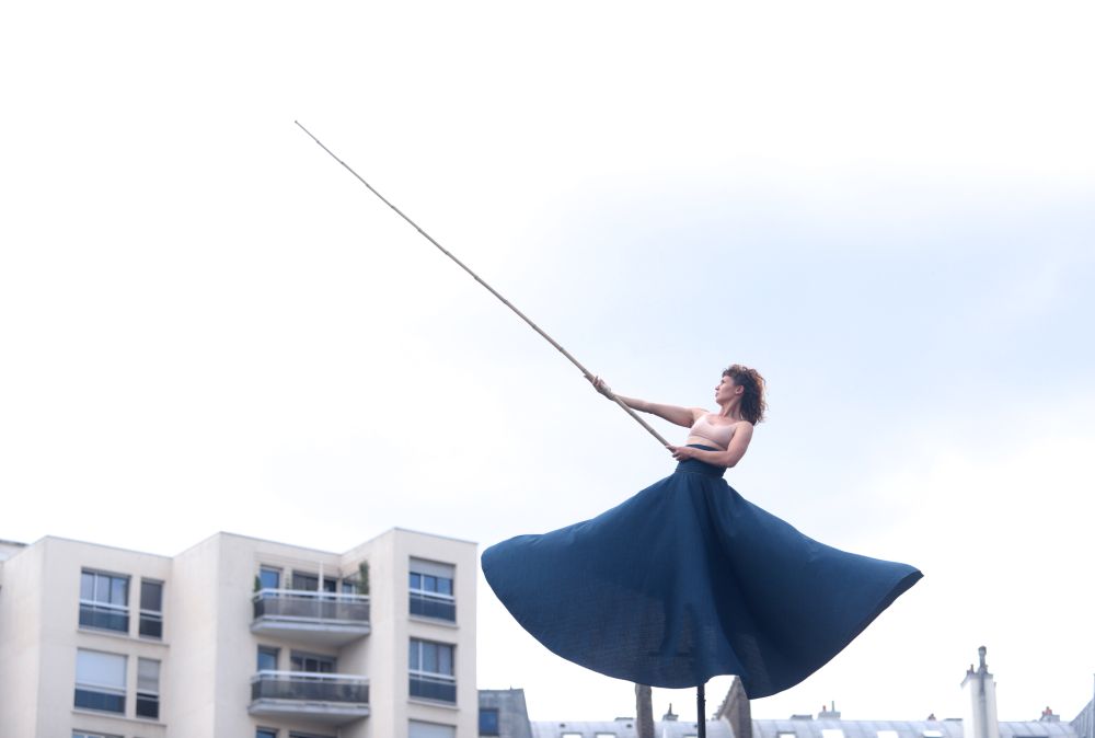 Kobieta w długiej rozkloszowanej spódnicy na wysokiej tyczce trzyma w rękach długi, prosty drąg. W tle blok mieszkalny i jasne niebo.