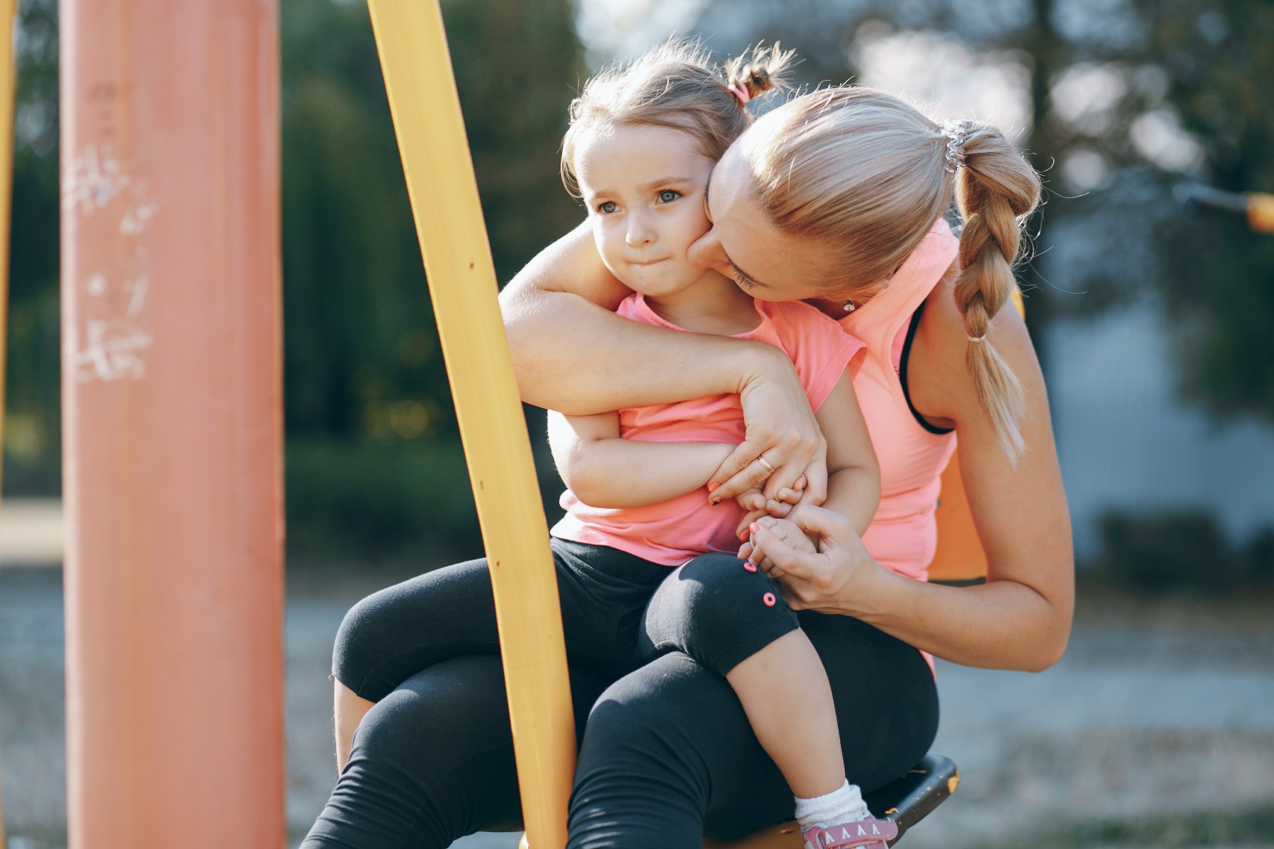 Kobieta w sportowym stroju siedzi na placu zabaw i czule obejmuje małą dziewczynkę w różowej koszulce, całując ją w policzek. Dziecko siedzi na jej kolanach, a w tle widać elementy konstrukcji placu zabaw oraz zieleń.