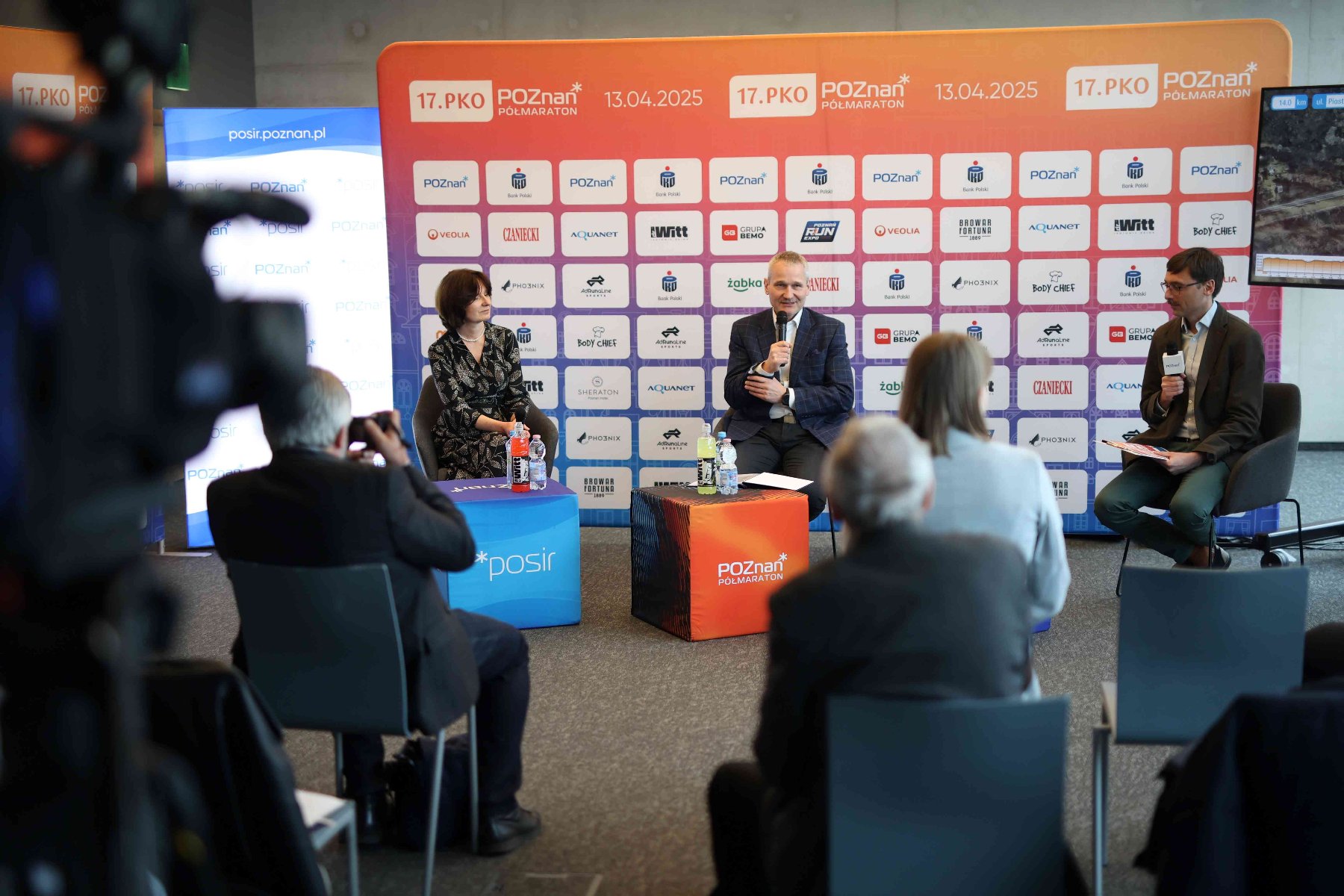 Konferencja przed 17. PKO Poznań Półmaratonem, fot. Studio Soho - na tle ścianki siedzą 3 osoby, 2 mężczyzn i kobieta. Mężczyzna w środku ubrany jest w marynarkę i mówi przez mikrofon. Mikrofon trzyma też mężczyzna z prawej strony, który również ma marynarkę. Z przodu widać kamerę i 3 osoby siedzące. Jeden siwy mężczyzna robi zdjęcie