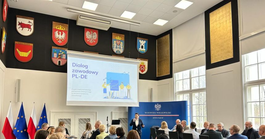 Konferencja w reprezentacyjnej sali urzędu z herbami województw na ścianie. Na ekranie widoczny tytuł prezentacji "Dialog zawodowy PL-DE'. Prowadząca stoi na tle niebieskiej ścianki z logo "Wojewoda Wielkopolski'. Wydarzenie dotyczy polsko-niemieckiej współpracy zawodowej, uczestnicy siedzą przy stołach konferencyjnych, przysłuchując się prezentacji.