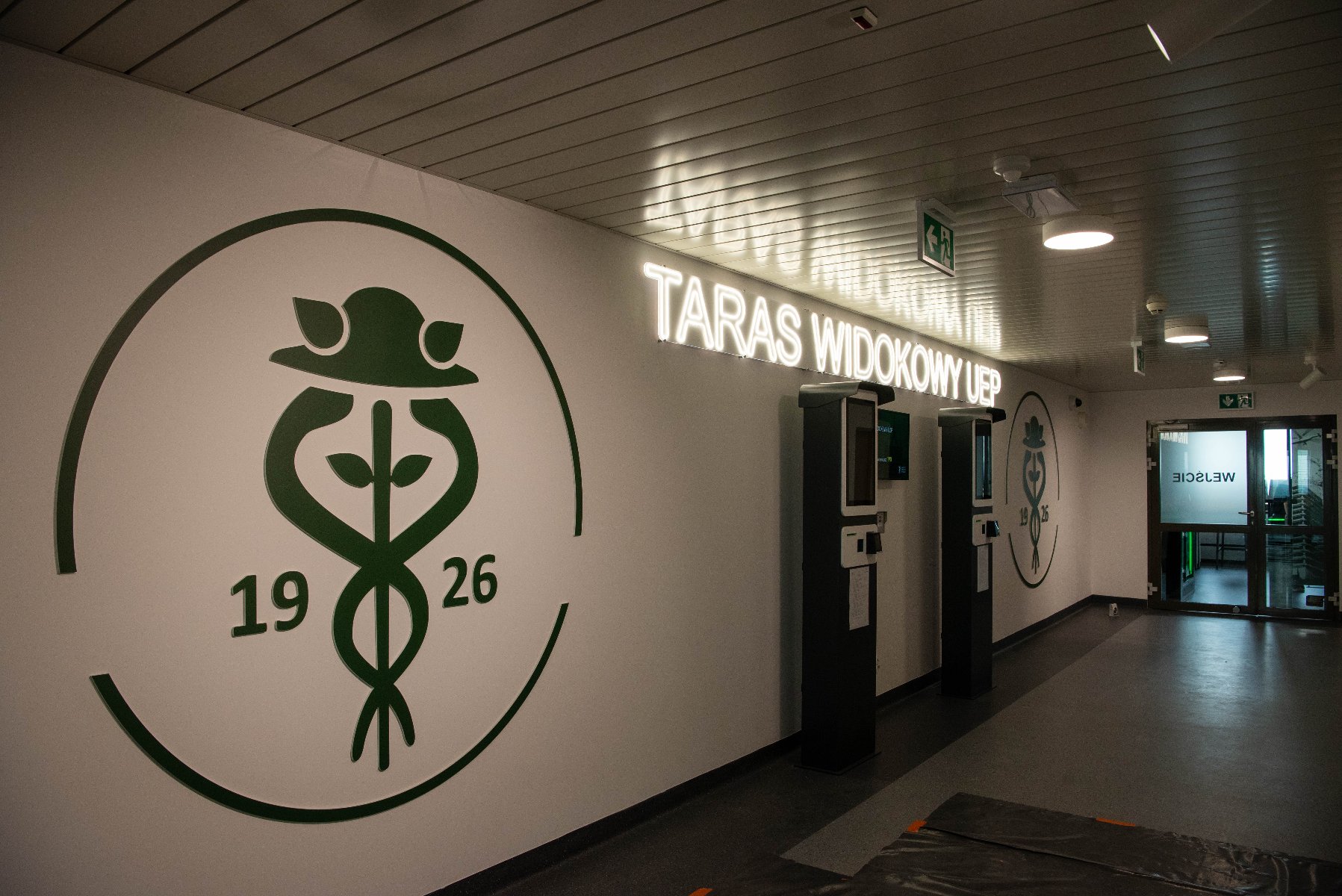 Korytarz z białymi ścianami. Na ścianie po lewej stronie znajduje się duże, zielone logo Uniwersytetu Ekonomicznego w Poznaniu z liczbami "19" i "26". Dalej, na ścianie, podświetlany napis "TARAS WIDOKOWY UEP". W oddali widoczne wejście do pomieszczenia.