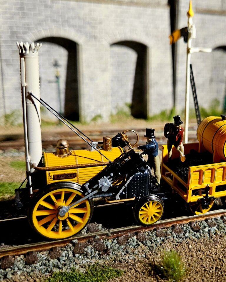 To jest miniaturowy model parowozu Rocket, historycznego pociągu, który zrewolucjonizował transport. Został ustawiony na makiecie torów kolejowych, kompletny z miniaturowym wagonem węglowym i figurkami ludzi. Model jest bardzo szczegółowy, z dokładnymi reprezentacjami projektu lokomotywy, w tym kotła, kół i nawet kominu. Model jest również pomalowany w jaskrawych kolorach, co dodaje realizmu.