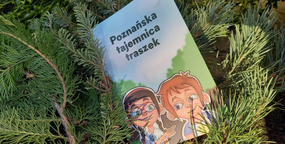 Książeczka dla dzieci zatytułowana "Poznańska tajemnica traszek" leży oparta o gałązki iglaków. Na okładce widnieje ilustracja dwójki uśmiechniętych dzieci - dziewczynki w okularach i chłopca - patrzących na małą traszkę trzymaną na dłoni.