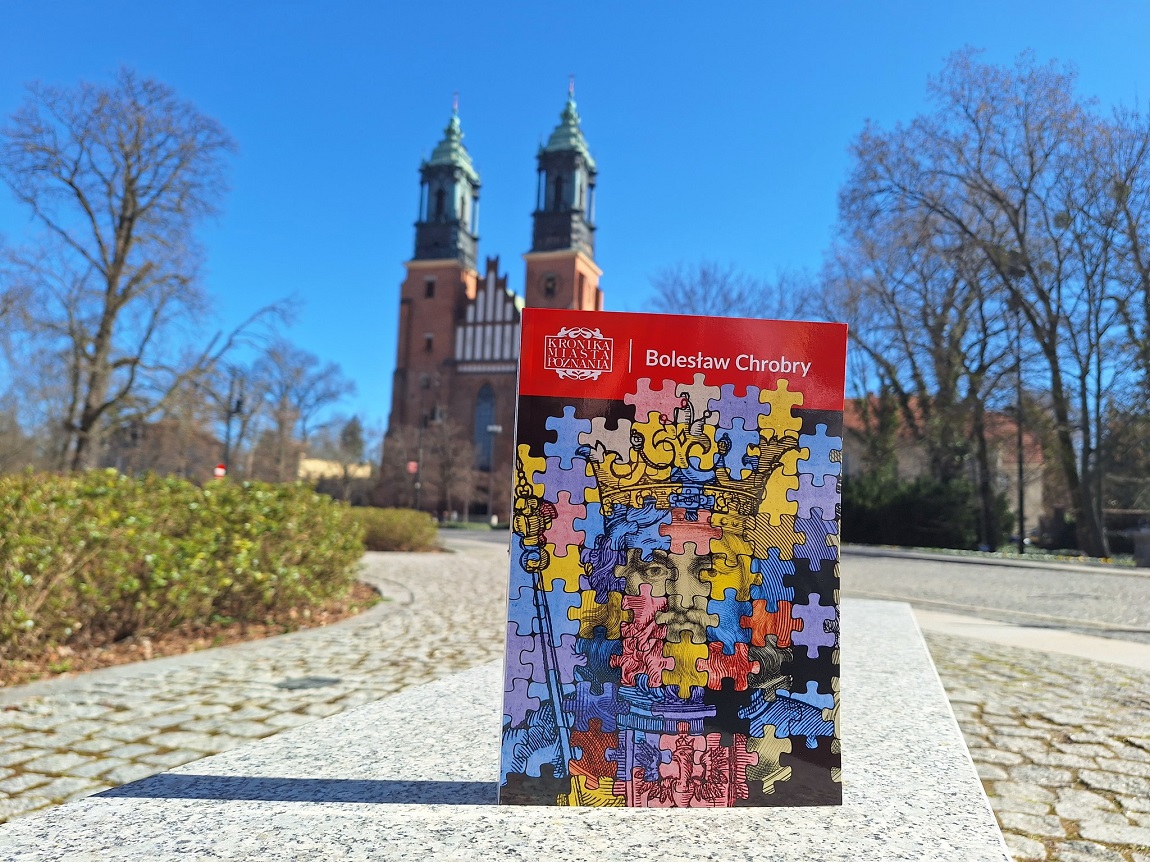 książka "Kronika Miasta Poznania Bolesław Chrobry" na okładce wizerunek króla z korona i zarostem, grafika stylizowana na układankę puzzle, książka stoi na kamiennym podłożu, w tle widoczna katedra poznańska