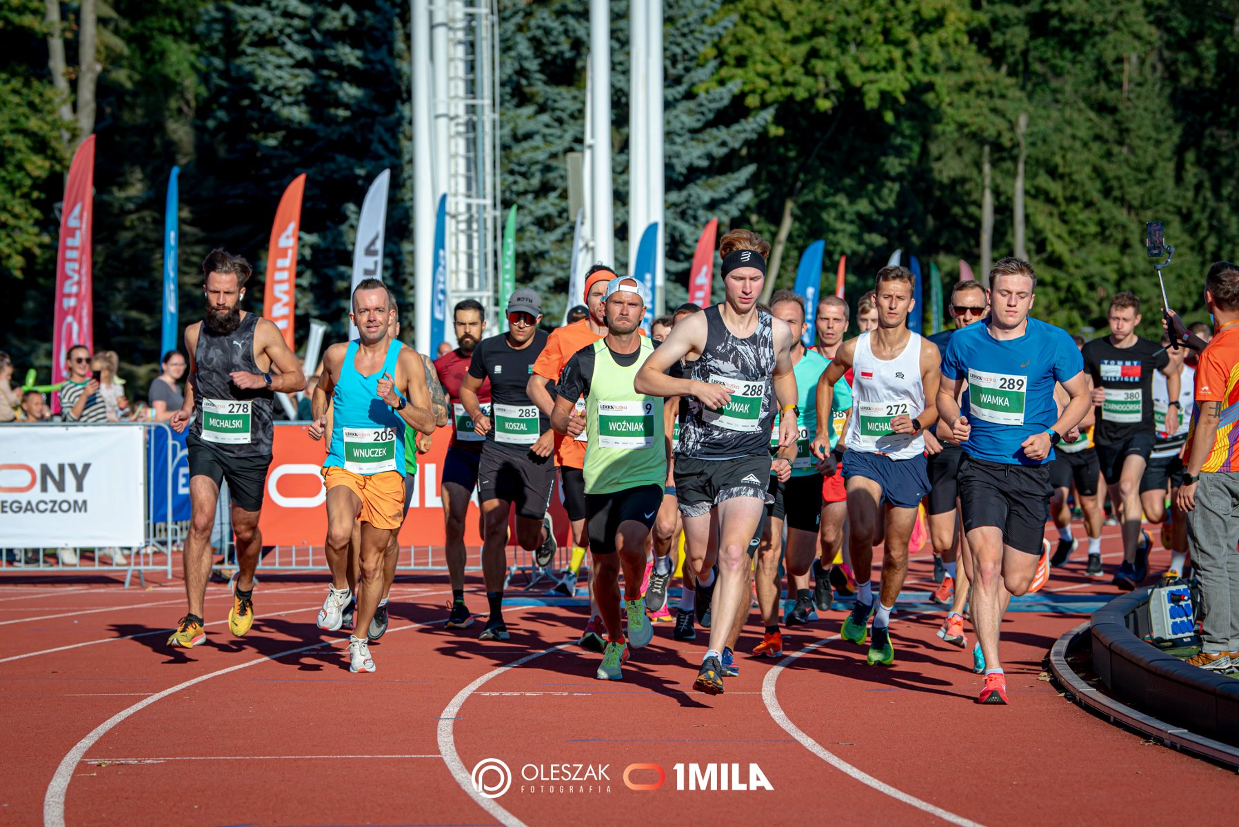 Zdjęcie, stadion, na którym biegnie grupa sportowców, za nimi flagi i drzewa.