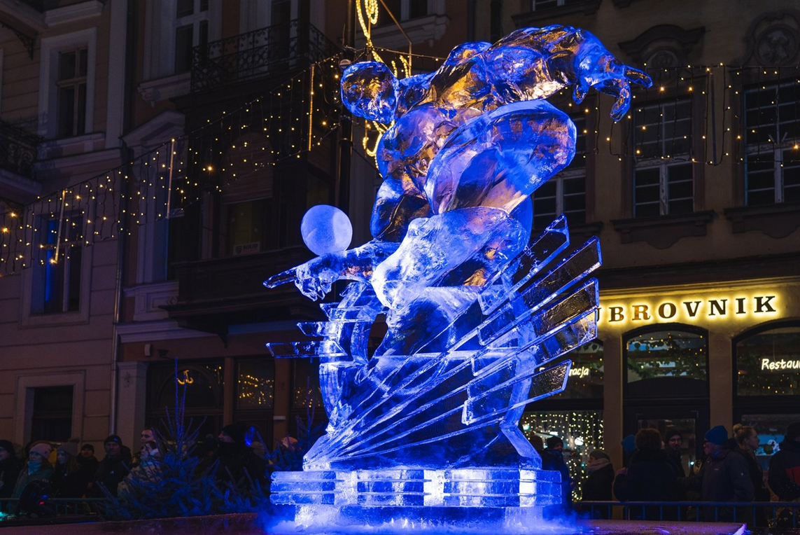 Lodowa rzeźba podświetlona na niebiesko. W tle świąteczne lampki oraz kamienice Starego Rynku w Poznaniu