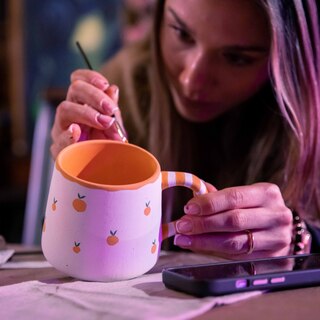 Kobieta koncentrującą się na ręcznym malowaniu ceramicznego kubka. Kubek ma biały kolor z pomarańczowym wnętrzem, a jego uchwyt jest ozdobiony paskami. Dodatkowo na powierzchni naczynia znajdują się małe, pomarańczowe owoce przypominające mandarynki.
