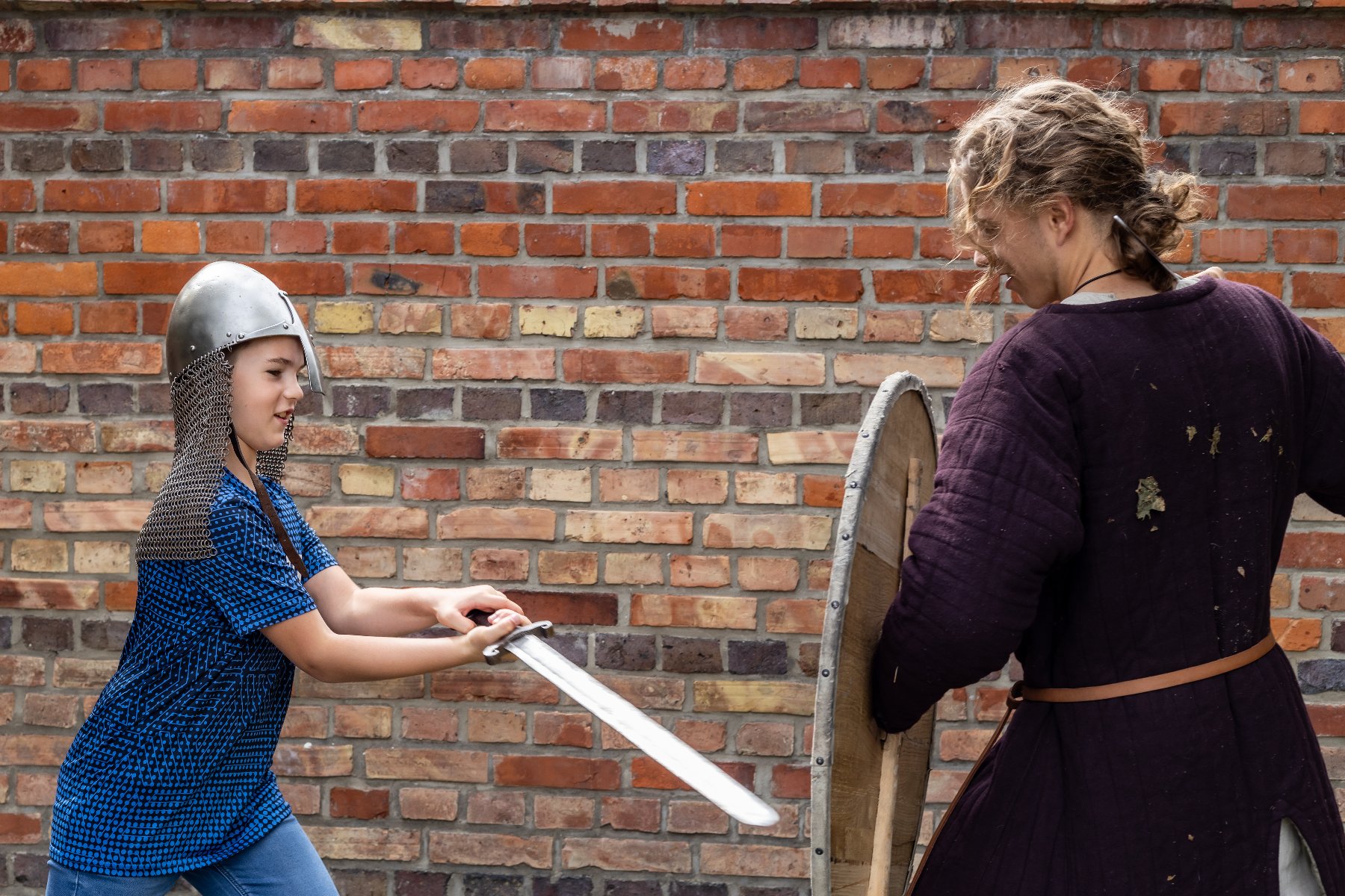 Mały chłopiec w T-shircie oraz w hełmie z mieczem. Obok mężczyzna z tarczą, ubrany w kostium. W tle ściana z cegły.