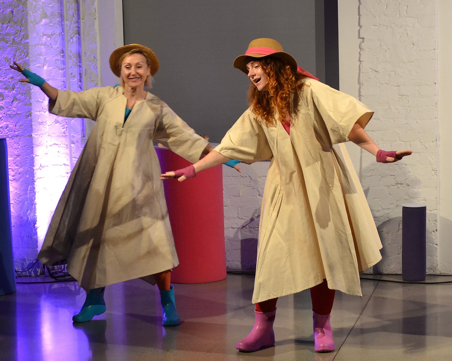 Na scenie dwie uśmiechnięte aktorki tańczą w pelerynach przeciwdeszczowych, kolorowych kaloszach i kapeluszach na głowie