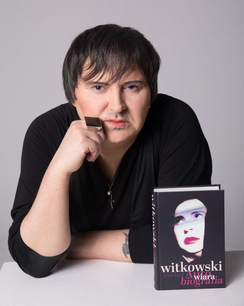 Mężczyzna ma opadającą na oczy grzywkę, na ręce duży pierścień z czarnym, kwadratowym kamieniem. Pozuje w czarnej koszuli obok książki "Wiara. Biografia".