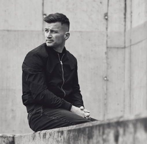 Mężczyzna w czarnej bluzie siedzi na murku, patrzy w dal.