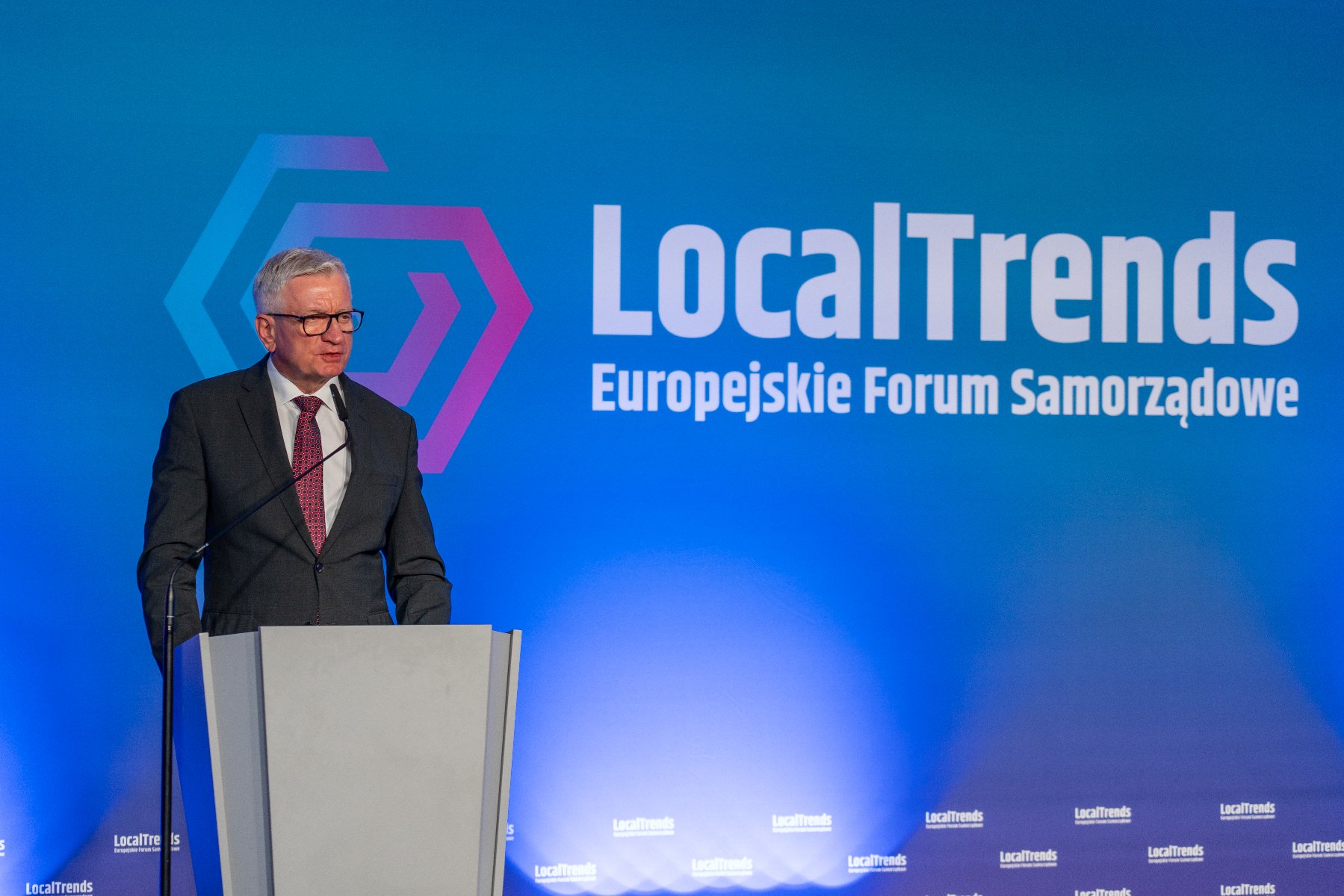 Mężczyzna w garniturze i okularach przemawia z mównicy. Za nim niebieskie tło z napisem "LocalTrends Europejskie Forum Samorządowe".