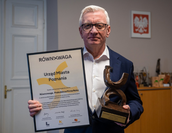 Mężczyzna w okularach i granatowej marynarce trzyma dyplom oraz brązową statuetkę. Na dyplomie napisy: "RÓWNOWAGA", "Urząd Miasta Poznania", "Równowaga. Prokobieca Firma Roku". W tle na ścianie godło Polski, po lewej białe drzwi.