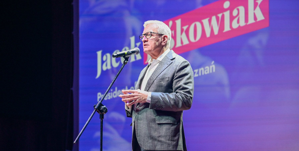 Mężczyzna w okularach i szarej marynarce stoi na scenie, przemawiając do mikrofonu. W tle fioletowy ekran z częściowo widocznym napisem: "Jacek Jaśkowiak" oraz "Prezydent Poznania".