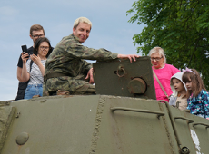 Mężczyzna w stroju żołnierza siedzi na czołgu. Uśmiecha się w stronę obiektywu. Obok niego stoją zwiedzający i przyglądają się pojazdowi.