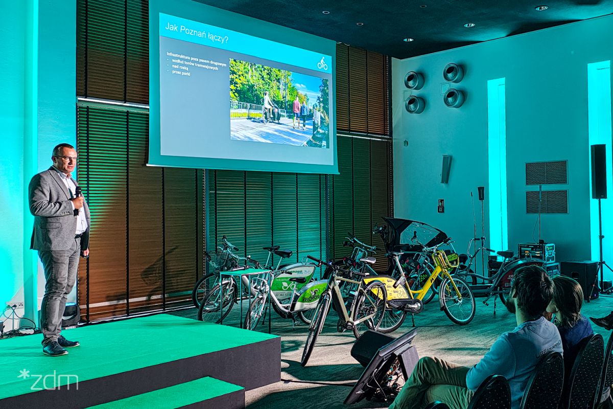 Mężczyzna w szarych spodniach, białej koszuli i szarej marynarce podczas wystąpienia na konferencji. Za nim ekran z wyświetlaną prezentacją, obok rozstawione rowery.