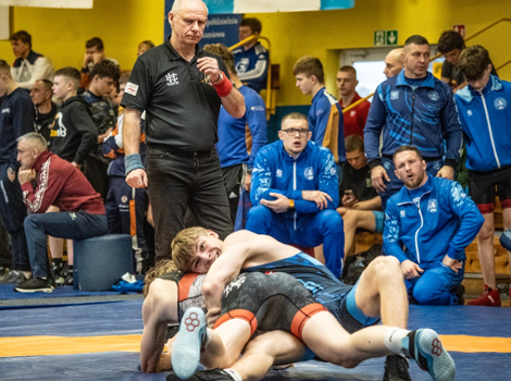 Międzynarodowy Turniej Zapaśniczy w Poznaniu, fot. Maciej Kula - 2 zawodnicy leżą na macie, w tle widać sędziego, przyglądających się trenerów oraz innych widzów na trybunach