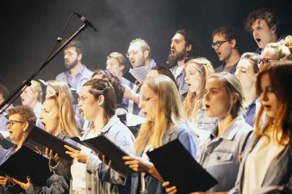Młoda dziewczyna mówi coś przez mikrofon, za nią chór.