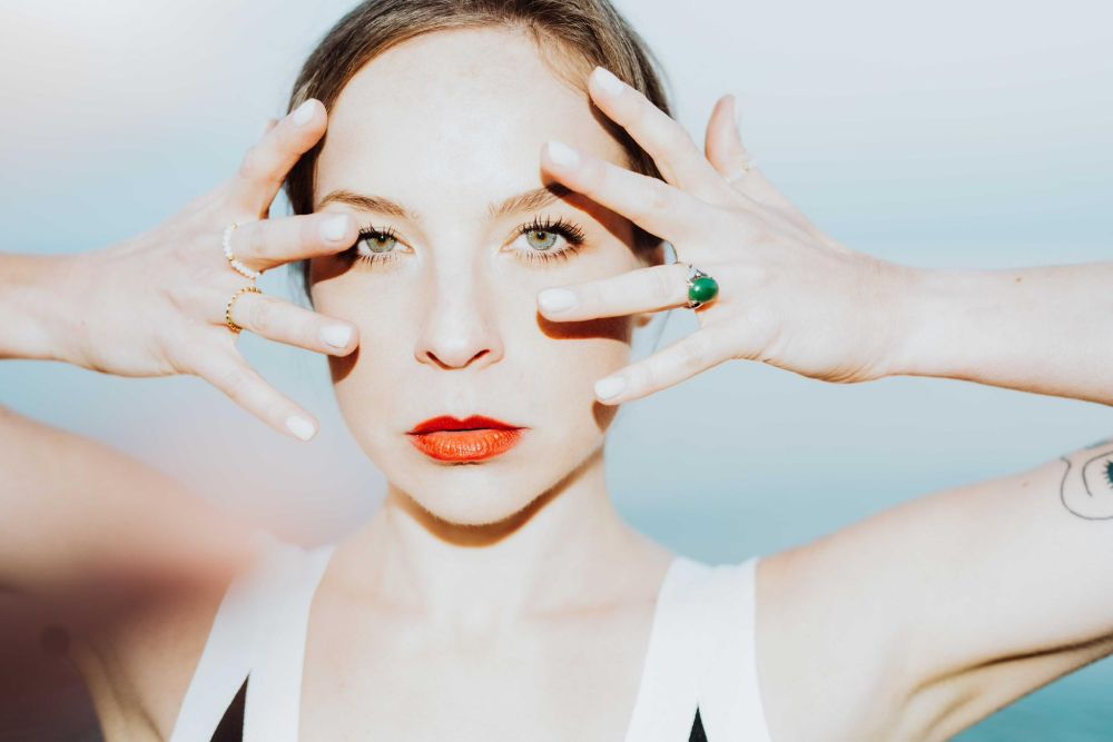 Młoda kobieta patrzy prosto w obiektyw muskając swoją twarz palcami na wysokości uszu.