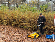 Młody mężczyzna w czarnej kurtce stoi w parku pełnym opadłych, brązowych liści. Obsługuje żółto-czarne urządzenie pomiarowe na kółkach (prawdopodobnie georadar lub skaner terenu) z przymocowanym tabletem.