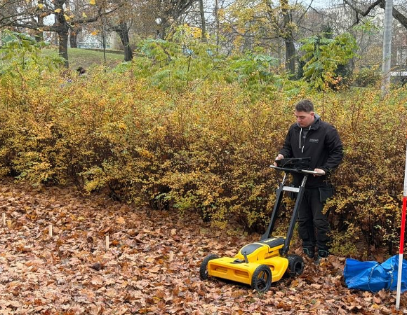 Młody mężczyzna w czarnej kurtce stoi w parku pełnym opadłych, brązowych liści. Obsługuje żółto-czarne urządzenie pomiarowe na kółkach (prawdopodobnie georadar lub skaner terenu) z przymocowanym tabletem.