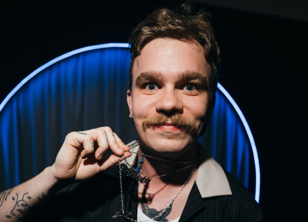 Młody mężczyzna z wąsem prezentuje swój wisiorek.