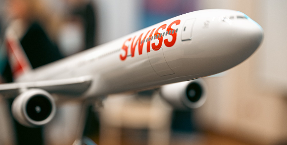 Model białego samolotu pasażerskiego na metalowym stojaku. Na kadłubie widoczny duży, czerwony napis "SWISS". Tło jest rozmyte, widać w nim zarysy postaci i wnętrza.