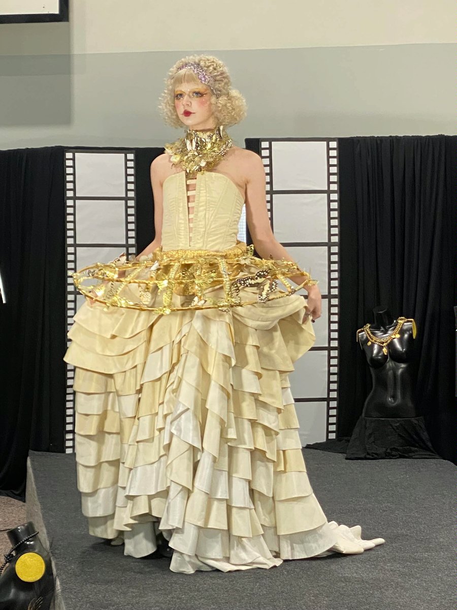 Modelka prezentuje spektakularną suknię na pokazie mody. Strój ma rozłożystą spódnicę z falbanami w kolorze kremowym oraz złote, błyszczące detale w górnej części. W tle dekoracja stylizowana na taśmę filmową oraz ekspozycja z ozdobami.