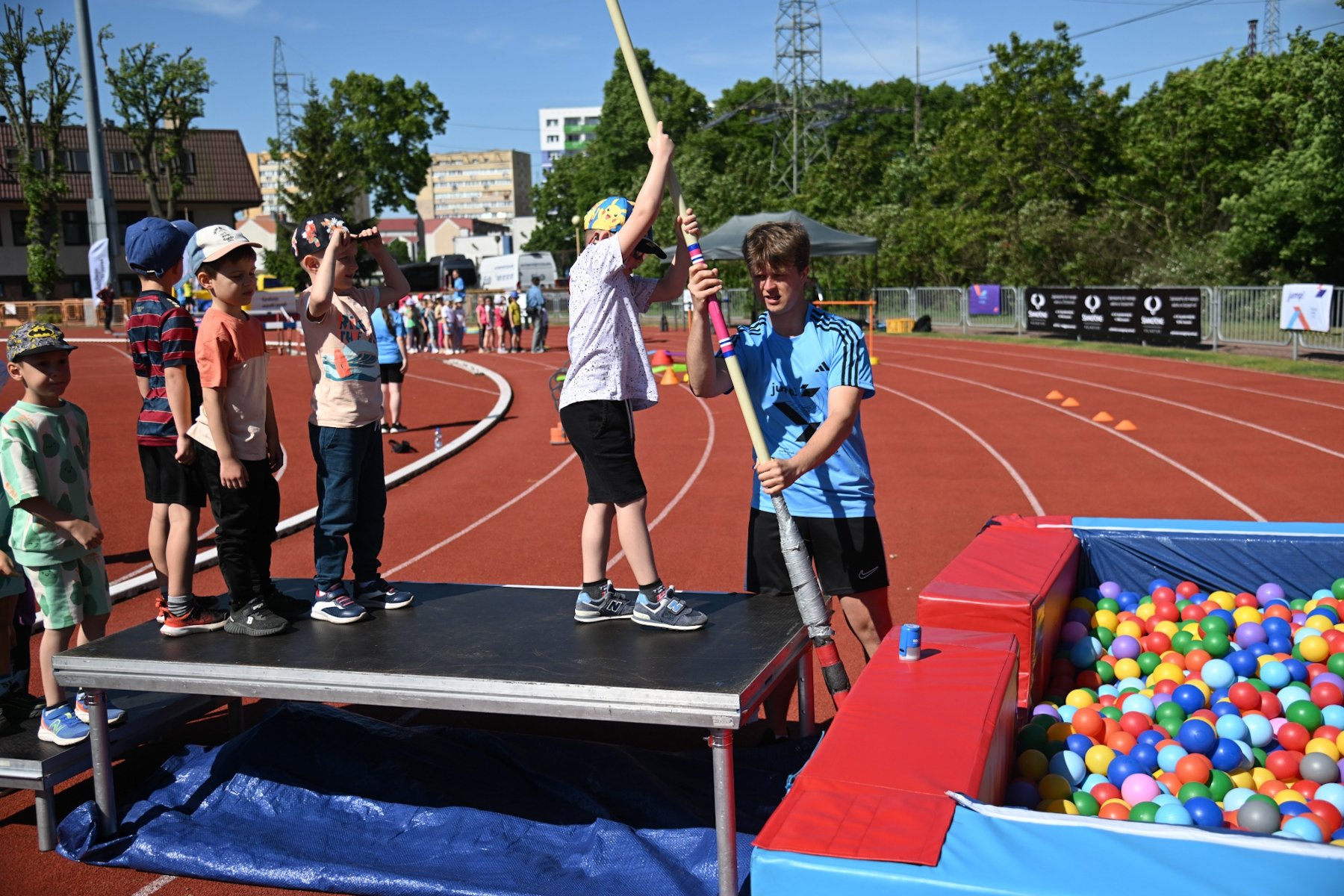 Monika Pyrek Tour, fot. Fundacja Moniki Pyrek - dzieci stoją na platformie i pierwsze z nich trzyma w ręce tyczkę. Przed nimi jest pojemnik z piłkami do ćwiczenia skoku