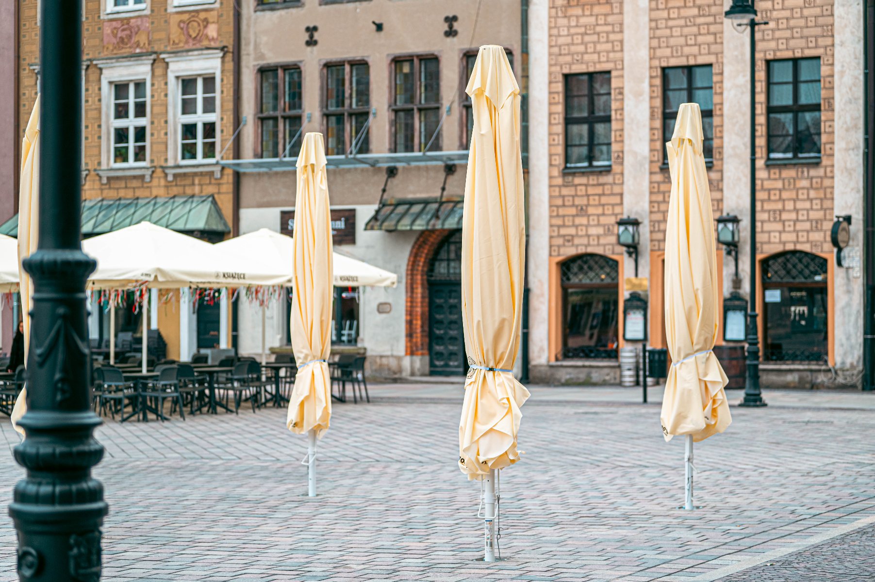 Na brukowanym placu stoją trzy złożone, wysokie parasole ogrodowe w kolorze kremowym. W tle, po lewej stronie, widać ogródek restauracyjny z rozłożonymi parasolami oraz czarnymi stolikami i krzesłami.