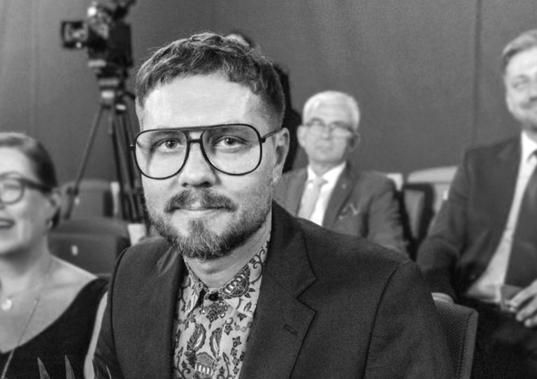 Na czarno-białym zdjęciu twarz mężczyzny w okularach, w tle inni ludzie