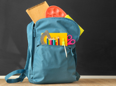 Na czarnym "tablicowm" tle znajduje się niebieski plecak z którego wystają przybory plastyczne, długopisy, kartki, ekierka, nożyczki itp.