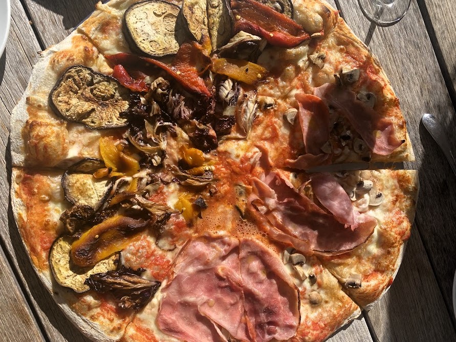 Na drewnianym stole leży pizza z różnymi dodatkami, szynka, bakłażan, papryka, pieczarki.