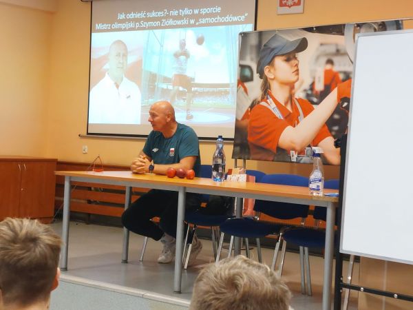 Na grafice przedstawiono mistrza olimpijskiego na spotkaniu z uczniami.