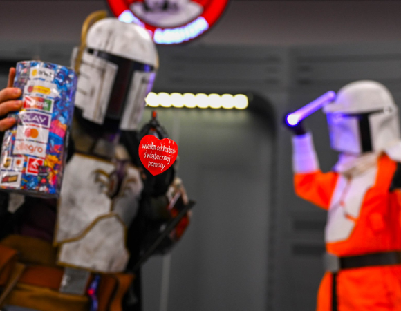 Na pierwszym planie widać czerwone serduszko WOŚP. W tle stoją dwie postaci w strojach z Gwiezdnych wojen