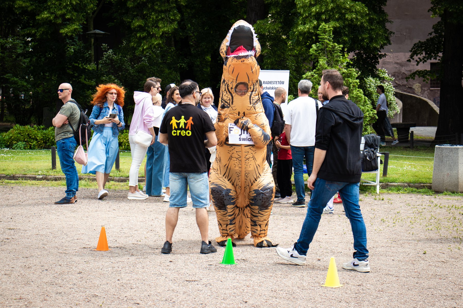 Na pierwszym planie widać osobę w stroju dinozaura i dwie osoby obrócone tyłem, a na drugim planie znajduje grupa osób.