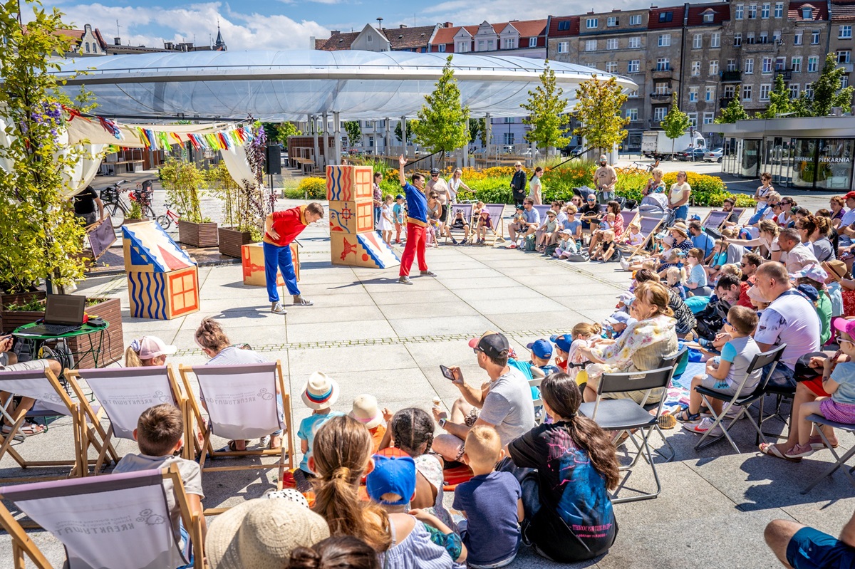 Na płycie Rynku Łazarskiego dwóch mężczyzn - aktorów występuje przed zgromadzoną publicznością. Jest to spekltakl dla dzieci. W tle widać kopułę zadaszenia rynku i kamienice.